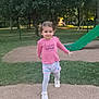 child, girl, playground, slide, grass, trees, park, pink_shirt, shorts, white_shoes, outdoor, smiling, young_child, fence, daylight, play_area, person, nature, walking, casual_clothing