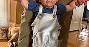 Brooks Bennett is registered to the contest to win money with this photo: toddler, child, adult, hands, tattoos, overalls, striped_clothing, blue_shirt, sneakers, wooden_floor, dog, indoor, standing, helping, person, casual_clothing, home, flooring, relaxed, cute