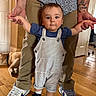 toddler, child, adult, hands, tattoos, overalls, striped_clothing, blue_shirt, sneakers, wooden_floor, dog, indoor, standing, helping, person, casual_clothing, home, flooring, relaxed, cute