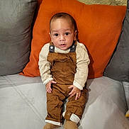 Naël participe au concours pour gagner de l'argent avec cette photo : baby, infant, child, couch, pillow, orange_pillow, gray_pillow, overalls, striped_shirt, socks, indoor, seated, curious, looking_at_camera, person, young_child, furniture, home, casual_clothing, portrait