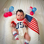 Enzo is registered to the contest to win money with this photo: baby, infant, flag, american_flag, balloons, patriotic, white_background, cushion, clothing, socks, person, indoor, celebration, cute, portrait, newborn, holiday, festive, red, blue