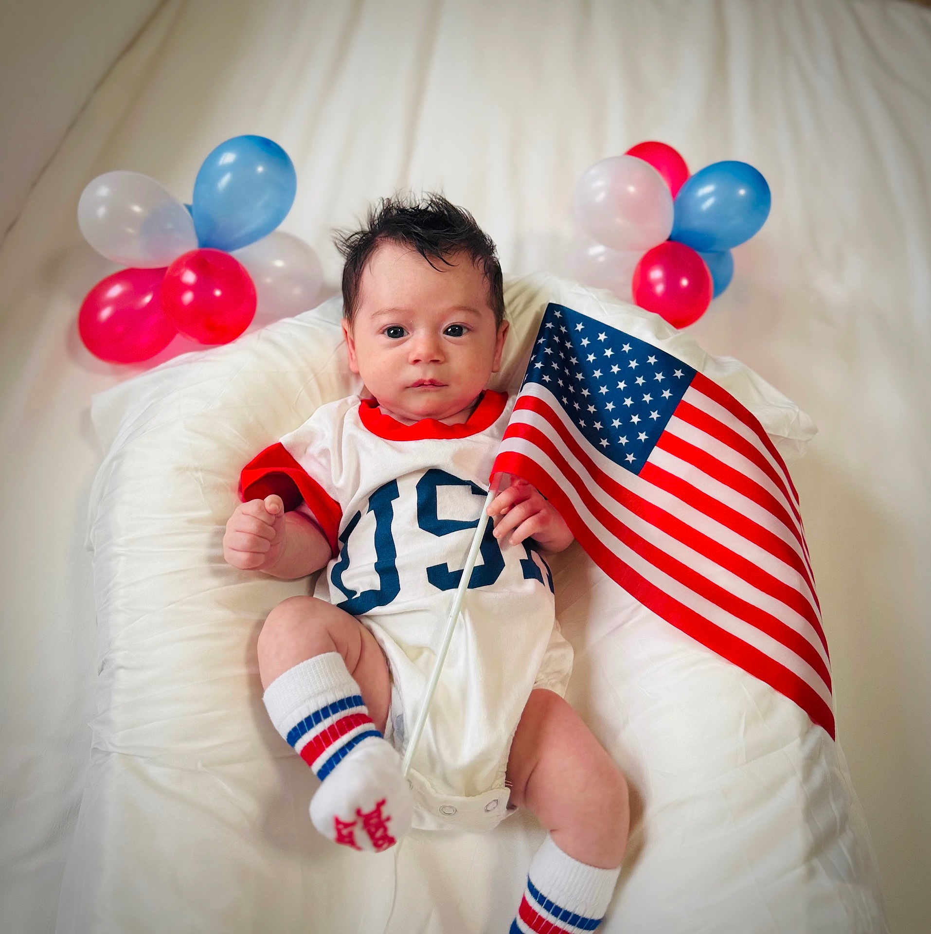 Enzo is registered to the contest to win money with this photo: baby, infant, flag, american_flag, balloons, patriotic, white_background, cushion, clothing, socks, person, indoor, celebration, cute, portrait, newborn, holiday, festive, red, blue