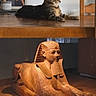 cat, tabby_cat, wooden_table, sphinx_statue, ancient_art, sculpture, indoor, furniture, chair, framed_photo, family, portrait, decor, relaxing_cat, animal, pet, stone, floor, room, lighting