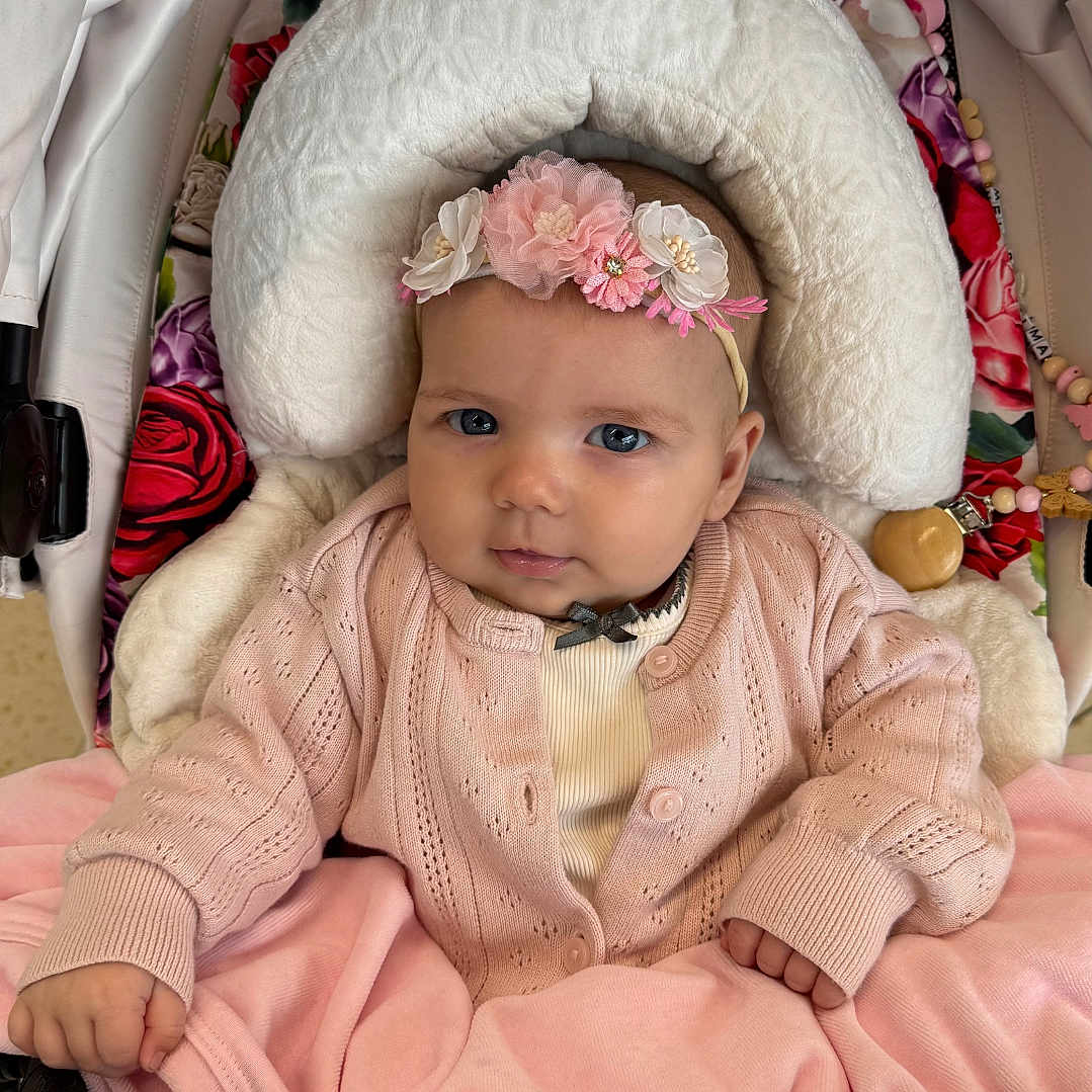 Melody is registered to the contest to win money with this photo: baby, blanket, child, closeup, comfort, cozy, cute, face, flower, hand, headband, indoors, infant, knitwear, pink_clothing, portrait, seated, soft_toys, stroller, young_child