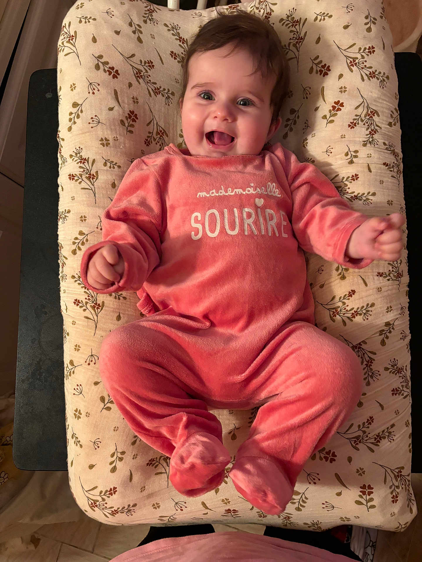 Garance participe au concours pour gagner de l'argent avec cette photo : baby, smiling, infant, pink_clothing, floral_pattern, cushion, happy, face, person, child, soft_texture, lying_down, indoors, cute, small_hands, footed_pajamas, joyful, cheerful, cozy, expression