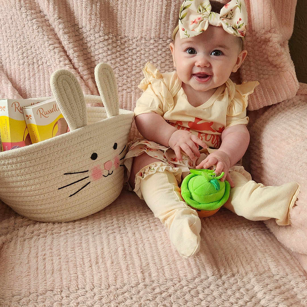 Eleanor is registered to the contest to win money with this photo: baby, child, infant, smiling, bow, yellow_clothing, soft_texture, pink_blanket, bunny_basket, toy, sitting, cute, happy, indoor, portrait, playful, cozy, decor, person, young_child