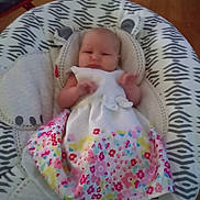 Hope is registered to the contest to win money with this photo: baby, infant, child, dress, floral_pattern, baby_seat, patterned_fabric, indoor, wood_floor, cute, young_child, person, small_hands, comfort, relaxing, home, seat_cushion, white_dress, colorful, adorable