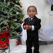 Symir joined the competition — help win amazing prizes! toddler, child, boy, suit, tie, christmas_tree, ornament, gift, present, holiday, festive, indoors, floor, decor, black_shoes, smiling, standing, white_blanket, red_bow, green_bow