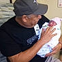 man, baby, newborn, hospital, cap, shirt, shorts, glasses, chair, blanket, person, indoor, holding, seated, tenderness, family, love, portrait, smile, hat