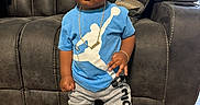 Shahaad is registered to the contest to win money with this photo: toddler, child, standing, blue_shirt, gray_pants, sneakers, gold_chain, curly_hair, living_room, couch, pillow, wooden_floor, indoor, person, young_child, casual_clothing, portrait, cute, fashion, home