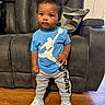 toddler, child, standing, blue_shirt, gray_pants, sneakers, gold_chain, curly_hair, living_room, couch, pillow, wooden_floor, indoor, person, young_child, casual_clothing, portrait, cute, fashion, home