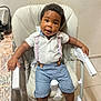 baby, child, high_chair, white_shirt, suspenders, blue_shorts, socks, indoor, curly_hair, looking_at_camera, furniture, table, floor_tiles, seat, cute, person, smiling, young, home, casual_clothing