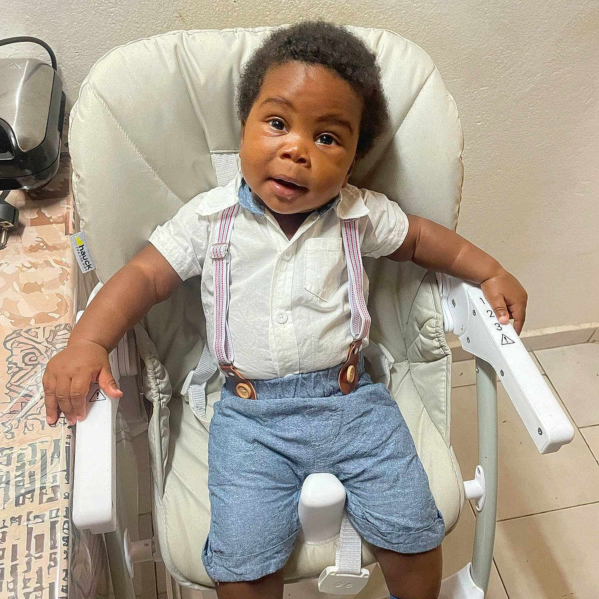 Jayce a rejoint le concours — aidez-le/la à gagner de superbes lots ! baby, blue_shorts, casual_clothing, child, curly_hair, cute, floor_tiles, furniture, high_chair, home, indoor, looking_at_camera, person, seat, smiling, socks, suspenders, table, white_shirt, young