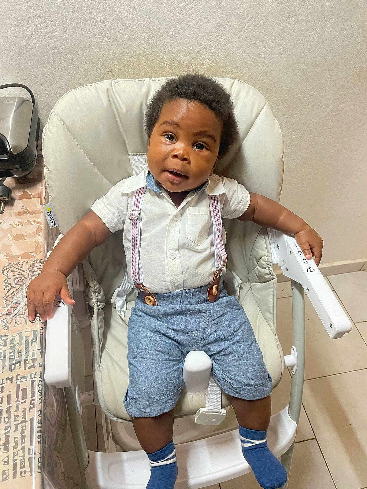 Jayce a rejoint le concours — aidez-le/la à gagner de superbes lots ! baby, child, high_chair, white_shirt, suspenders, blue_shorts, socks, indoor, curly_hair, looking_at_camera, furniture, table, floor_tiles, seat, cute, person, smiling, young, home, casual_clothing