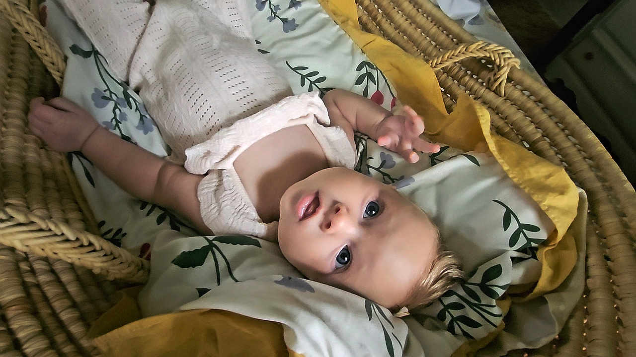 Myra is registered to the contest to win money with this photo: baby, infant, basket, blanket, floral_pattern, mustard_color, sleepwear, lying_down, curious, face, head, skin, hand, indoors, soft_texture, cozy, person, child, newborn, cute