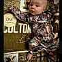 baby, infant, one_month, camouflage_clothing, blanket, animal_print, cow, wooden_sign, milestone, lying_down, curious_expression, newborn, child, portrait, cute, indoors, soft_texture, clothing, face, hand