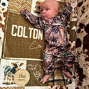 Colton is registered to the contest to win money with this photo: baby, infant, one_month, blanket, cow_pattern, wooden_sign, camouflage_clothing, lying_down, newborn, feet, hand, face, head, plaid, animal_print, soft_texture, cute, milestone, portrait, indoor