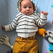 Noa a rejoint le concours — aidez-le/la à gagner de superbes lots ! baby, child, striped_hoodie, mustard_pants, socks, blue_background, soft_toys, teething_toy, rattle, stuffed_animal, lying_down, infant, cute, curious_expression, indoors, playtime, colorful_toys, soft_texture, casual_clothing, baby_fashion