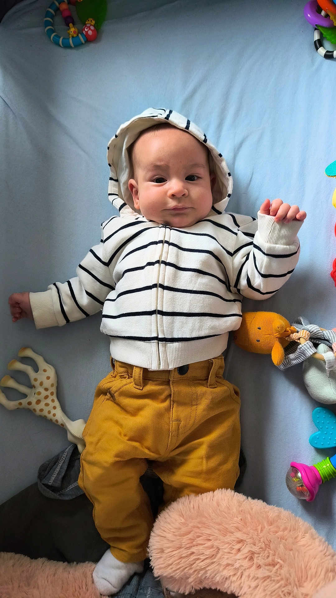 Noa a rejoint le concours — aidez-le/la à gagner de superbes lots ! baby, child, striped_hoodie, mustard_pants, socks, blue_background, soft_toys, teething_toy, rattle, stuffed_animal, lying_down, infant, cute, curious_expression, indoors, playtime, colorful_toys, soft_texture, casual_clothing, baby_fashion