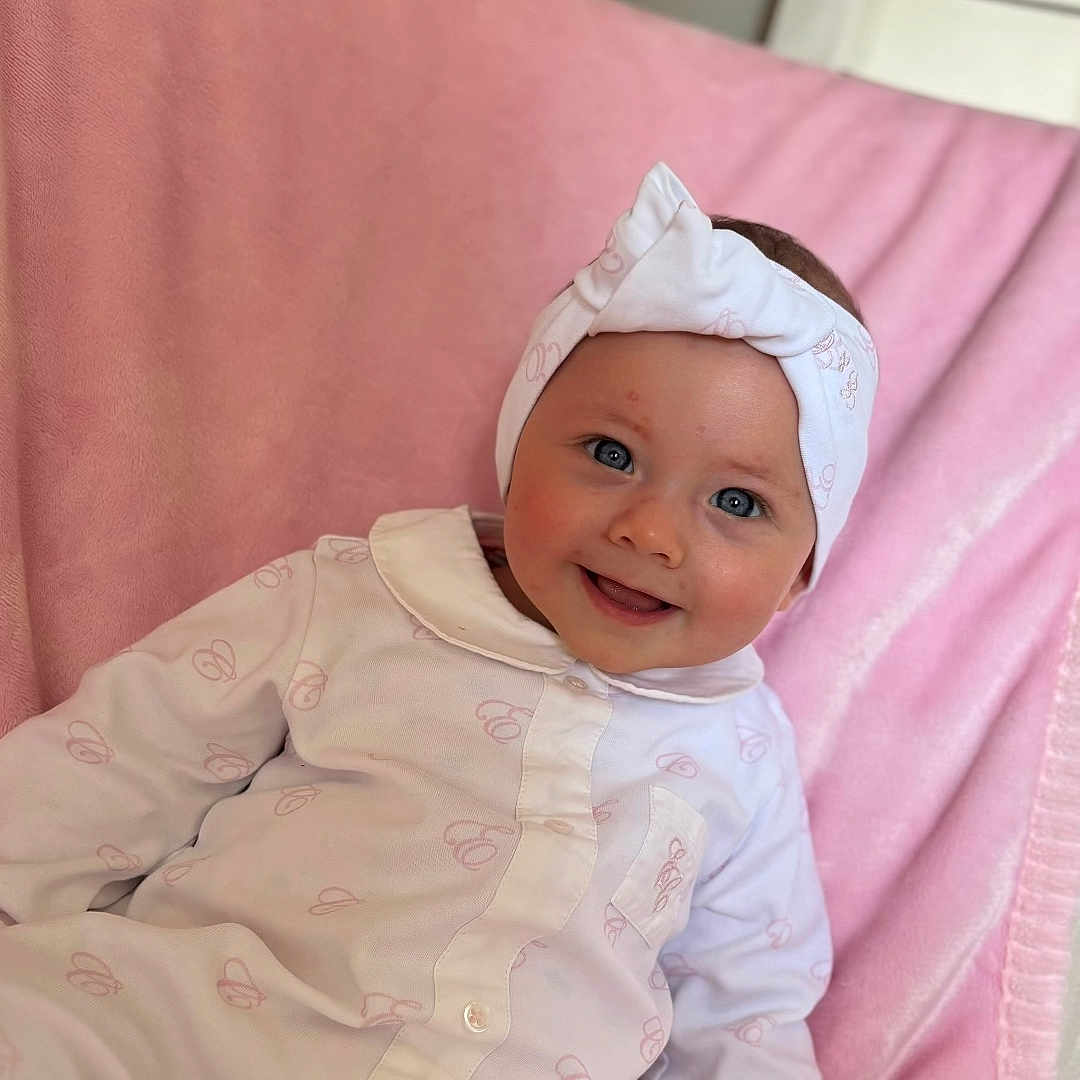 Djynn a rejoint le concours — aidez-le/la à gagner de superbes lots ! baby, baby_girl, child, closeup, clothing, comfortable, cozy, cute, face, happy, headband, indoor, infant, onesie, person, pink_blanket, portrait, sitting, smiling, soft_texture