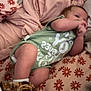 baby, infant, child, bed, pillow, blanket, onesie, shoes, footwear, sleeping_area, floral_pattern, fabric, resting, cute, indoor, soft, comfort, person, laying_down, hand
