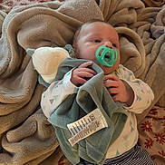 Maxwell is registered to the contest to win money with this photo: baby, pacifier, blanket, plush_toy, bedspread, infant, clothing, striped_pants, patterned_shirt, soft_texture, bedroom, sleeping_area, comfort, cute, hand, face, head, lying_down, resting, warm