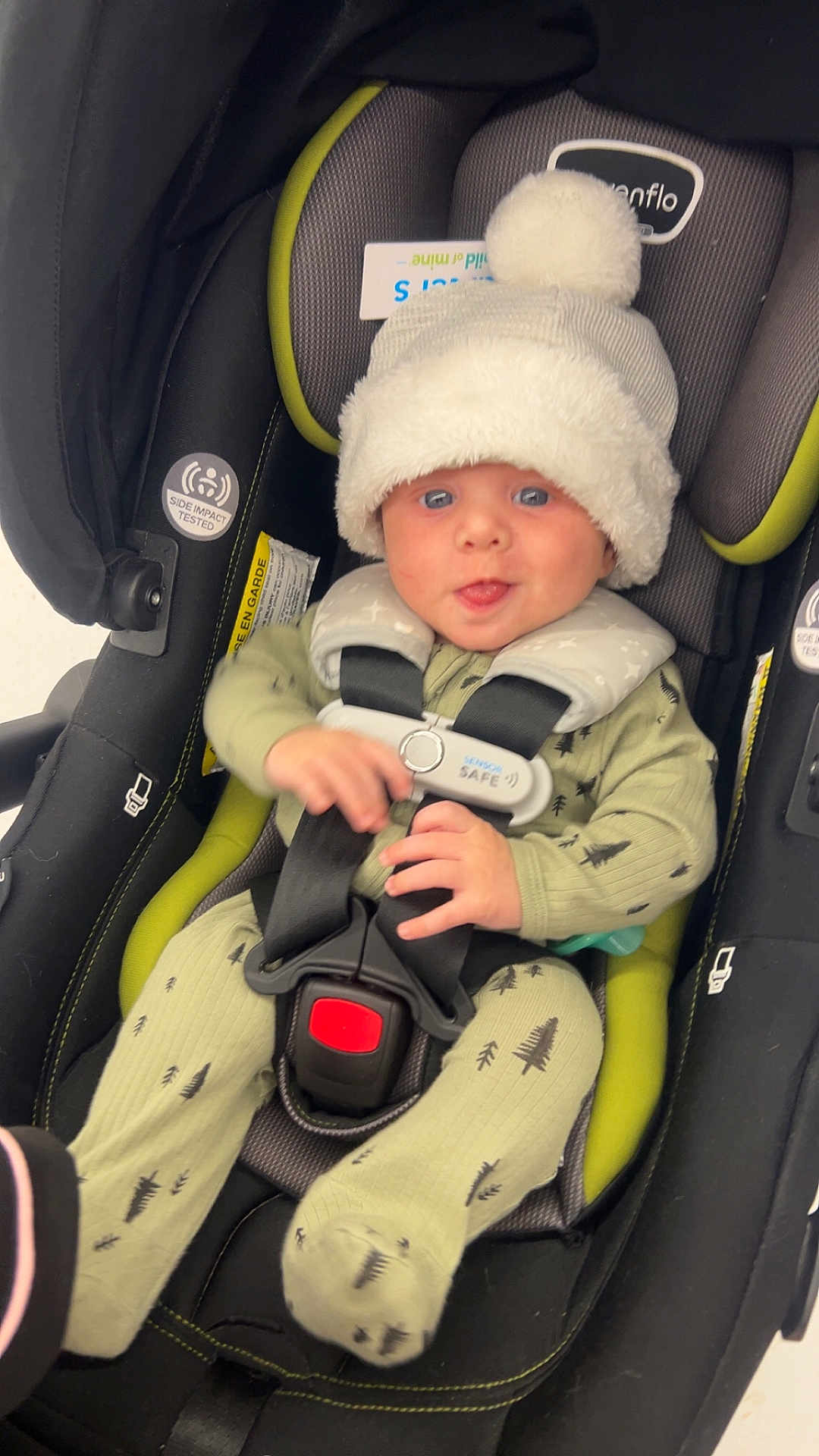 Maxwell is registered to the contest to win money with this photo: baby, infant, car_seat, hat, pom_pom, pajamas, green_clothing, tree_pattern, smiling, tongue_out, child, seat_belt, safety_harness, indoor, cute, cozy, happy, person, childcare, transportation