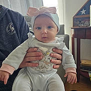 Raelynn joined the competition — help win amazing prizes! baby, infant, headband, bow, clothing, person, adult_hands, couch, indoor, flag_display_case, elephant_cartoon, text_on_clothing, cozy, portrait, child, cute, holding, soft_lighting, room, furniture