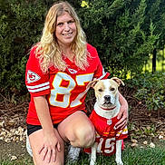 Tommy is registered to the contest to win money with this photo: woman, person, dog, pet, outdoor, garden, grass, greenery, portrait, smiling, kneeling, football_jersey, sports, team, red_clothing, matching_outfits, collar, shorts, nature, photography