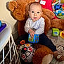 baby, teddy_bear, toy_cube, soccer_ball, plush_toy, play_area, child, indoor, floor_mat, socks, long_sleeve_shirt, pants, colorful_toys, soft_toy, curious, seated, wide_eyes, light, home, cute