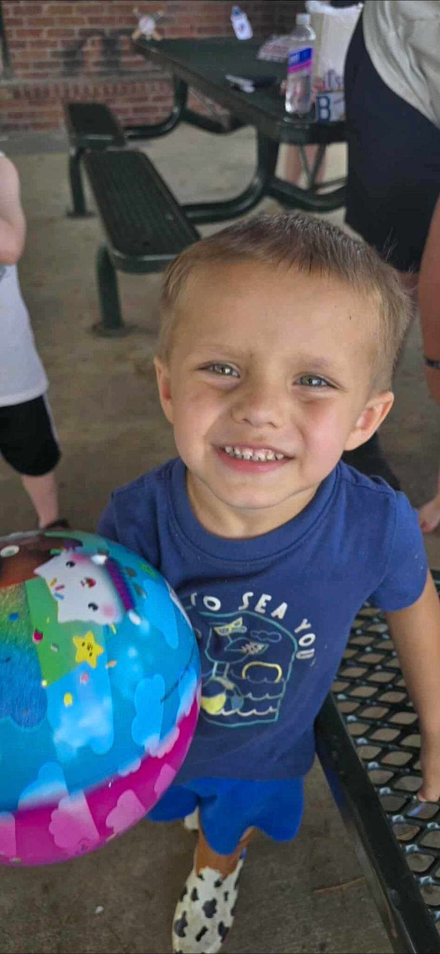 Wyatt is registered to the contest to win money with this photo: child, smiling, ball, blue_shirt, shorts, outdoor, picnic_table, brick_wall, person, happy, toy, footwear, concrete_floor, daylight, casual_clothing, playful, close_up, young_child, holding_object, background_people