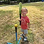 child, boy, smiling, baseball_bat, tee_ball, yellow_ball, grass, field, shorts, tshirt, sneakers, outdoor, daylight, trees, sports, play, recreation, happy, standing, sunny