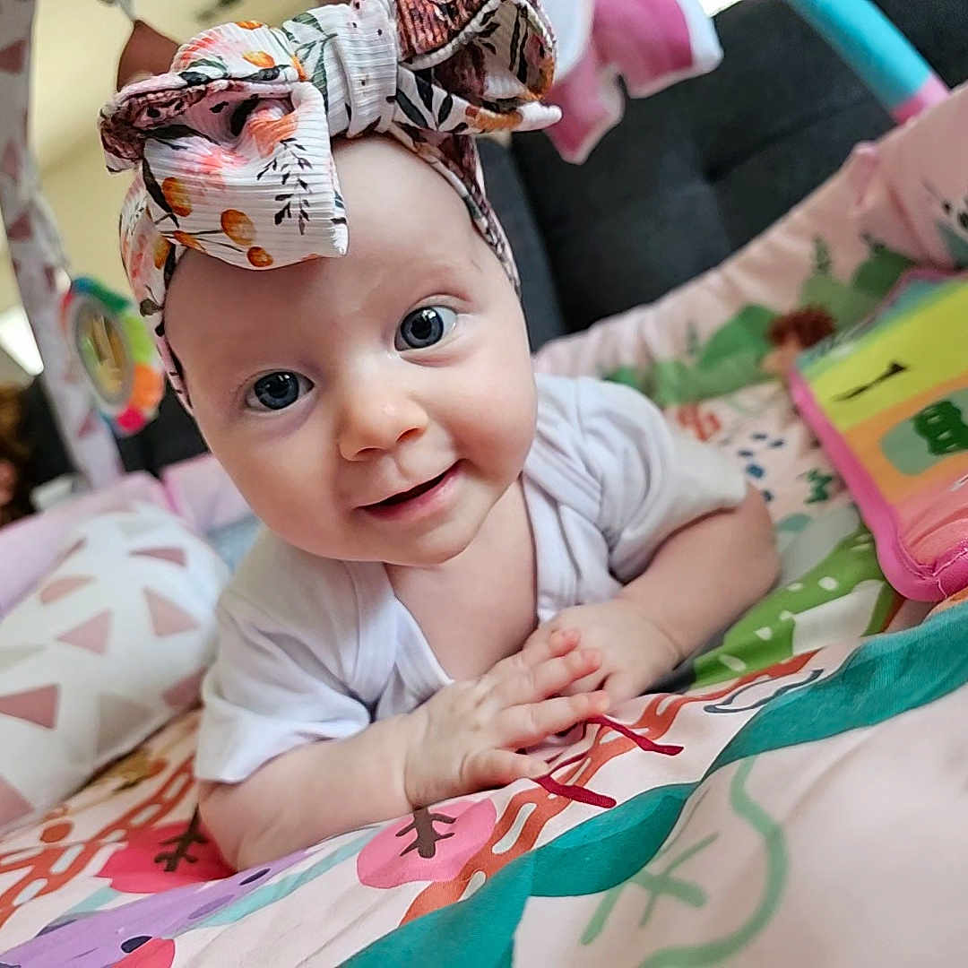 Charlotte is registered to the contest to win money with this photo: baby, background, blurred, candid, child, clothing, colorful, curious, cute, face, hands, headband, indoor, infant, lying_down, person, play_mat, smiling, soft_toys, young_child