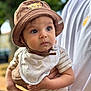 baby, child, hat, brown_hat, lion_cub_design, bib, striped_clothing, onesie, person, adult, hand, outdoor, greenery, portrait, curious, alert, holding, closeup, soft_light, background_blur