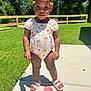 toddler, child, outdoor, sunlight, grass, fence, concrete, romper, sandals, sippy_cup, hair_beads, serious_expression, standing, daytime, nature, trees, shoes, person, kid, playtime