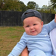 Jett is registered to the contest to win money with this photo: baby, child, infant, portrait, smiling, happy, cap, backwards_cap, blue_clothing, outdoor, backyard, grass, fence, trees, stroller, arms, feet, sitting, family, daylight