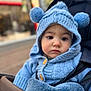 baby, blue_clothing, buttoned_sweater, child, close_up, cute, face, infant, knit_hat, outdoor, pom_poms, portrait, seated, serious_expression, soft_texture, stroller, urban_background, warm_clothing, winter_wear, young_child