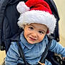 Alexander is registered to the contest to win money with this photo: child, toddler, santa_hat, smiling, face, blue_clothing, stroller, baby, cute, holiday, festive, winter, young_child, portrait, happy, outdoor, seatbelt, hat, person, seasonal