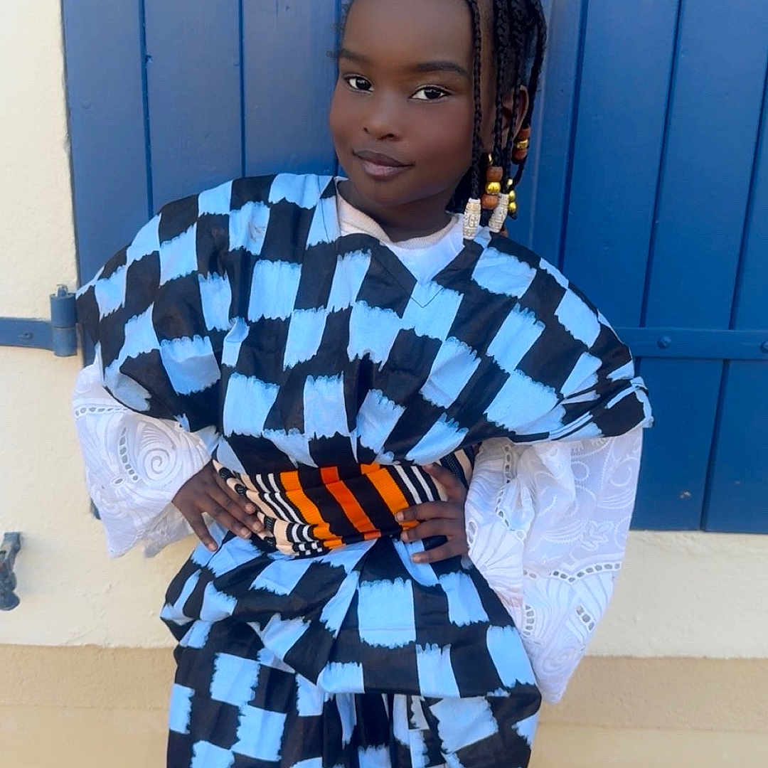 Amsatou participe au concours pour gagner de l'argent avec cette photo : child, portrait, outdoor, fashion, clothing, patterned_outfit, blue, black, orange, window, shutter, wall, standing, posing, confident, beaded_hair, smiling, person, youth, street