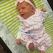 Marcellina is registered to the contest to win money with this photo: baby, bed, child, cozy, cute, face, foot, hand, headband, indoor, infant, lying_down, newborn, patterned_blanket, person, pink_clothing, sleepwear, smiling, soft_toys, striped_blanket