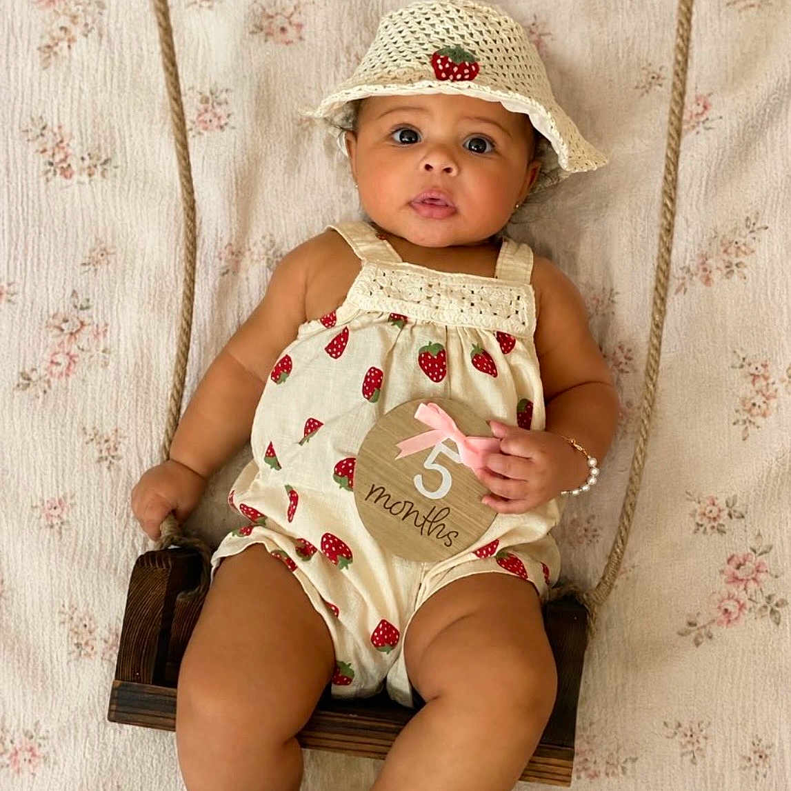 Ariyah is registered to the contest to win money with this photo: baby, infant, child, portrait, sitting, swing, sun_hat, strawberry_pattern, romper, floral_background, wooden_swing, milestone_photo, five_months, facial_expression, dark_hair, brown_eyes, indoors, fashion, cute, photography