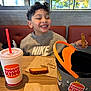 child, smiling, curly_hair, nike_sweatshirt, burger_king, fast_food, french_fries, drink_cup, table, napkin, restaurant_interior, window, daylight, food, happy, casual_clothing, indoor, kid, eating, seat