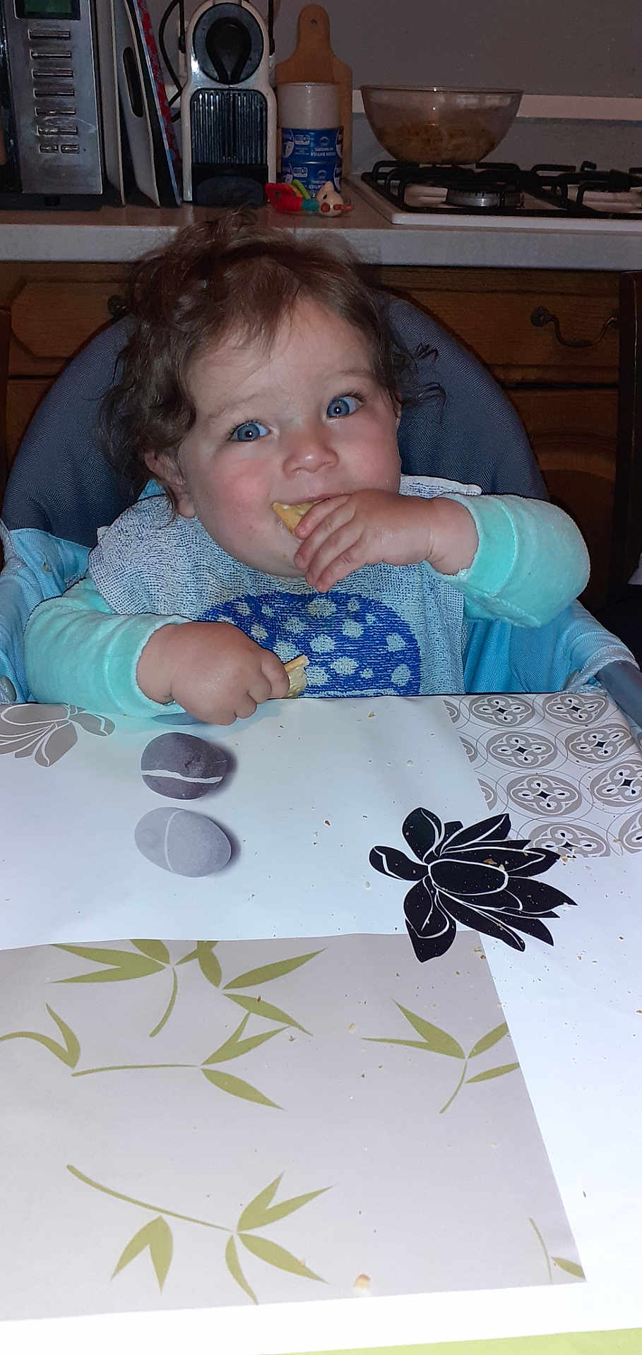 Noah a rejoint le concours — aidez-le/la à gagner de superbes lots ! toddler, child, high_chair, snack, blue_eyes, curly_hair, kitchen, table, bib, eating, food, countertop, stove, utensils, plate, person, indoor, happy, cute, baby