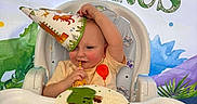 Kaleb is registered to the contest to win money with this photo: baby, child, birthday, birthday_cake, high_chair, party_hat, dinosaur, dinosaur_theme, smiling, happy, sitting, indoor, celebration, food, cake, colorful, portrait, infant, festive, playful