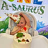 baby, child, birthday, birthday_cake, high_chair, party_hat, dinosaur, dinosaur_theme, smiling, happy, sitting, indoor, celebration, food, cake, colorful, portrait, infant, festive, playful