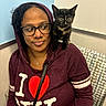 black_cat, casual_clothing, cat, chair, cozy, curly_hair, expression, glasses, hoodie, indoor, jewelry, kitten, looking_away, necklace, pattern, portrait, seated, shoulder, tortoiseshell_cat, woman