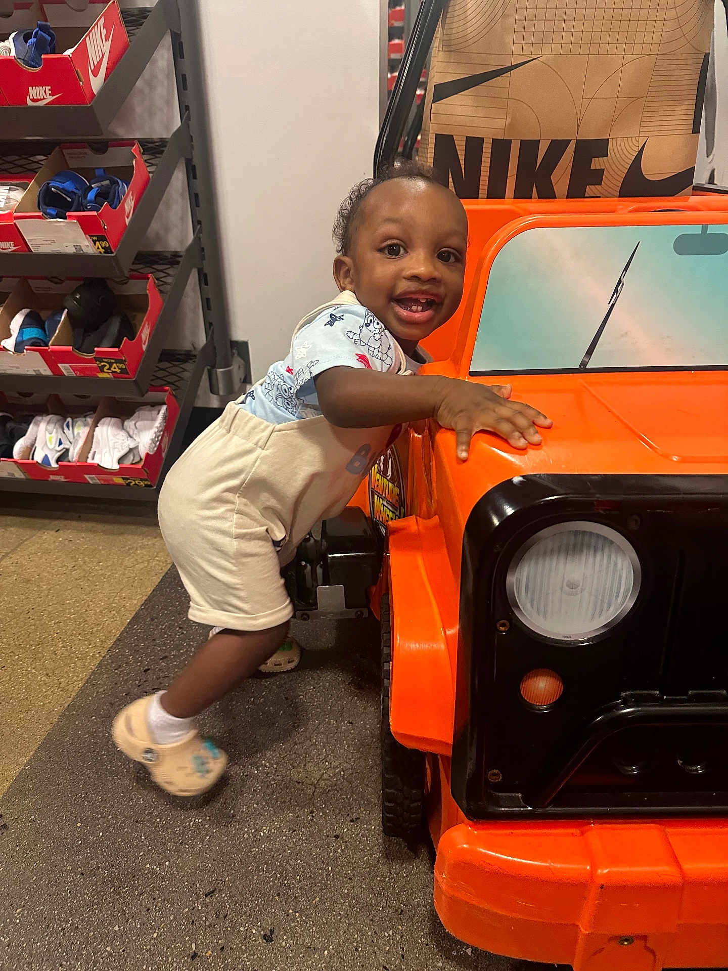 Vincent is registered to the contest to win money with this photo: toddler, child, toy_car, orange, smiling, indoor, shoes, nike, shelf, footwear, clothing, white_socks, play, happy, person, floor, retail_store, curly_hair, cute, fun