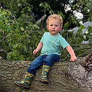 Kaiden is registered to the contest to win money with this photo: boots, casual_clothing, child, curly_hair, daylight, green, jeans, leaves, nature, outdoor, playful, portrait, rain_boots, serious_expression, sitting, toddler, tree, trunk, tshirt, young_child