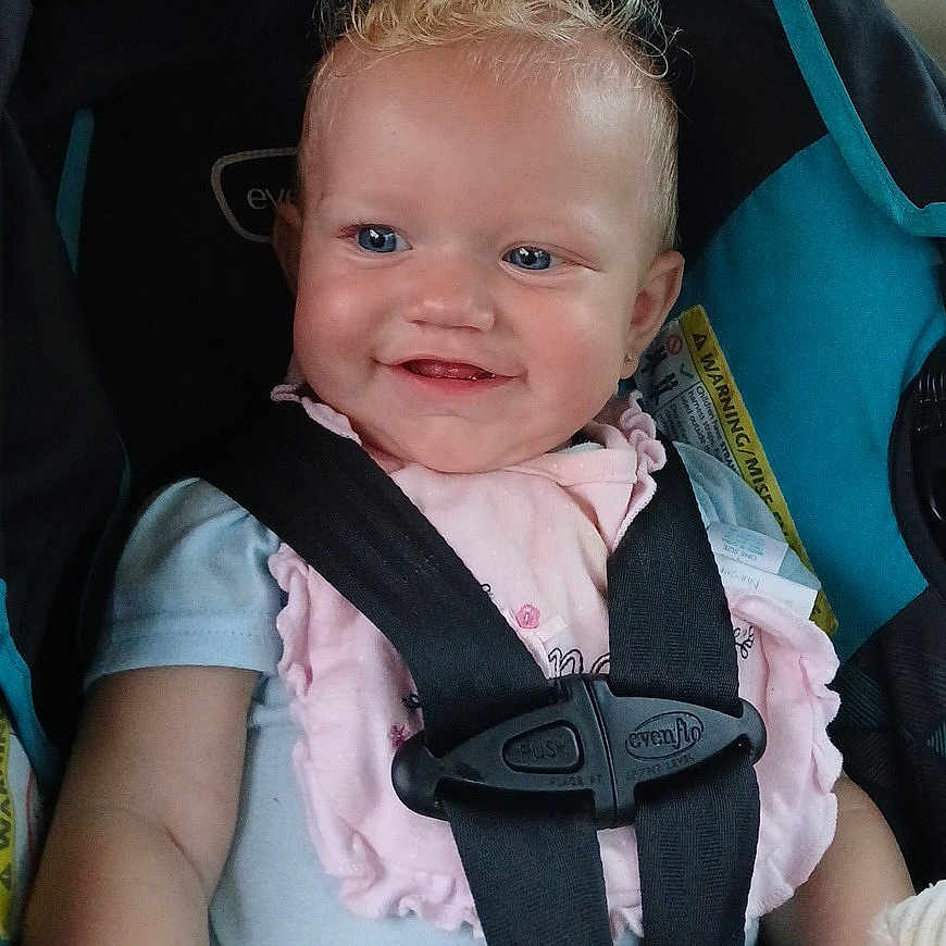 Arayah is registered to the contest to win money with this photo: baby, blonde, blue_eyes, car_seat, child, clothing, curly_hair, cute, happy, indoors, infant, person, pink_bib, portrait, safety_harness, seat, seatbelt, secure, smiling, young_child