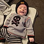 baby, infant, smiling, hat, beanie, sweater, skull, crossbones, striped_pants, couch, blanket, mat, indoor, child, clothing, happy, person, cute, portrait, resting